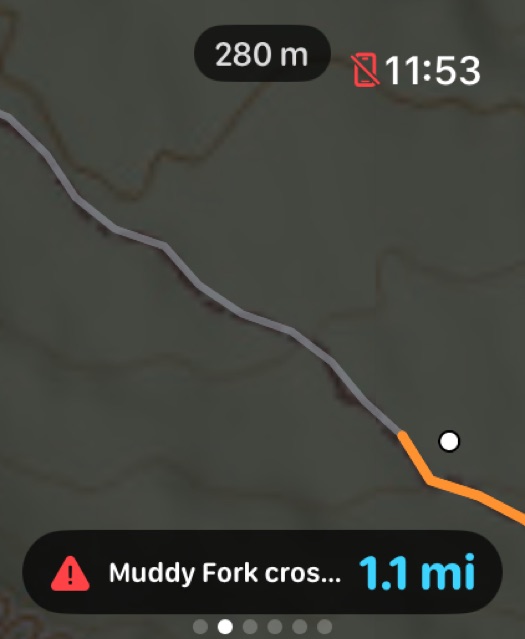 Apple Watch screen showing the trail zoomed in, the walked section in grey and the trail ahead in orange, a white dot marking position, a 280 metre scale pill, and a red hazard row reading Muddy Fork crossing 1.1 miles.